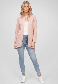 Lichtroze langarm cardigan met een open voorkant, gecombineerd met een witte top en lichtblauwe high-waisted jeans, gedragen met witte sneakers.
