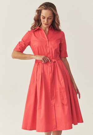 Femme ajustant la ceinture nouée à la taille de sa robe corail à boutons, longueur genou, avec des manches mi-longues.