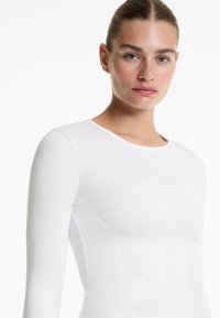 Top alb cu maneci lungi, din material texturat cu nervuri, cu decolteu rotund. Designul ajustat pune în evidență forma corpului prin detalii subtile de cusătură.
