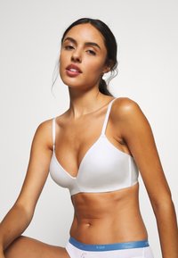 Soutien-gorge blanc sans coutures avec un design triangulaire, des bretelles réglables et une texture douce. Porté avec un sous-vêtement blanc présentant une ceinture bleue.