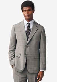 Giacca da completo a quadri grigia con risvolti a lancia, due bottoni e tasche laterali. Indossata sopra una camicia bianca con pois blu navy e una cravatta a righe.