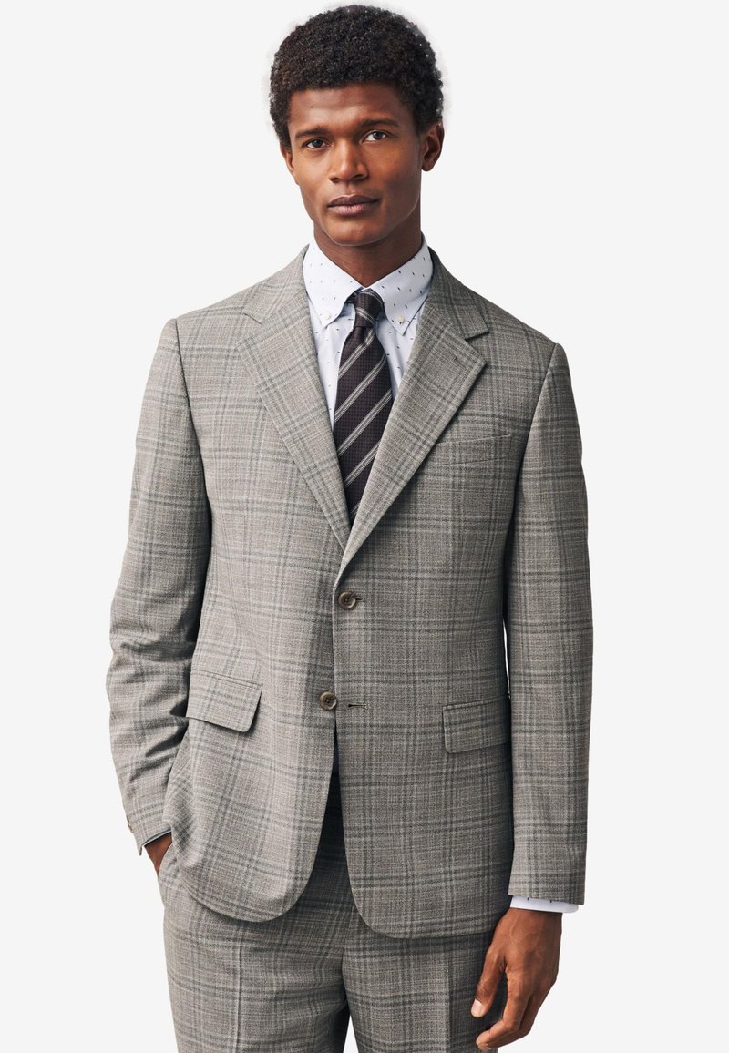 Giacca da completo a quadri grigia con risvolti a lancia, due bottoni e tasche laterali. Indossata sopra una camicia bianca con pois blu navy e una cravatta a righe.