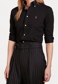 Femme portant une chemise noire boutonnée aux manches retroussées et un pantalon noir à fines rayures taille haute avec une ceinture assortie.