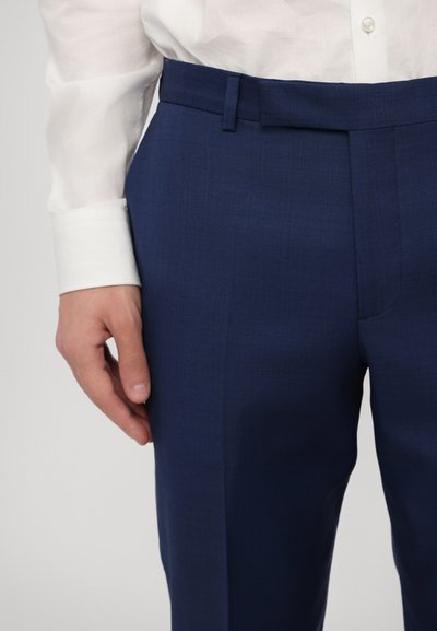 Pantalons de cérémonie bleu marine en tissu texturé, dotés d'une coupe ajustée, de passants de ceinture et d'une taille lisse, associés à une chemise blanche au niveau des poignets.
