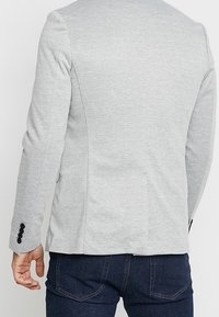 Hombre con una chaqueta gris claro con botones negros en las mangas y jeans azul oscuro, visto desde atrás contra un fondo liso.