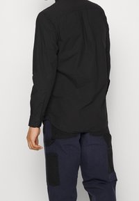 Maglietta nera a maniche lunghe con polsini a bottone e orlo curvo. Indossata sopra pantaloni cargo blu scuro, che mettono in evidenza dettagli di tessuto a contrasto.