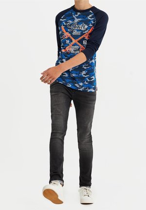 Blauwe camouflage langemouwen shirt met marineblauwe mouwen, voorzien van skategraphics in oranje en wit, gecombineerd met zwarte slim-fit jeans en witte sneakers.