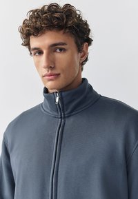 Grijze jas met hoge kraag en rits, gemaakt van zacht materiaal met een gladde textuur. Voorzien van een centrale rits en subtiele stiksel details.