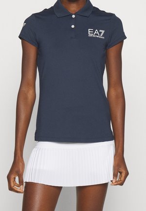 Vrouw draagt een marineblauw poloshirt met EA7 Emporio Armani-logo en een witte geplooide minirok, staand voor een effen achtergrond.