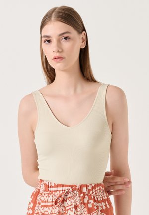 Canottiera a coste di un beige chiaro con scollatura a V. La modella l'ha abbinata a una gonna arancione a fantasia con disegni bianchi.