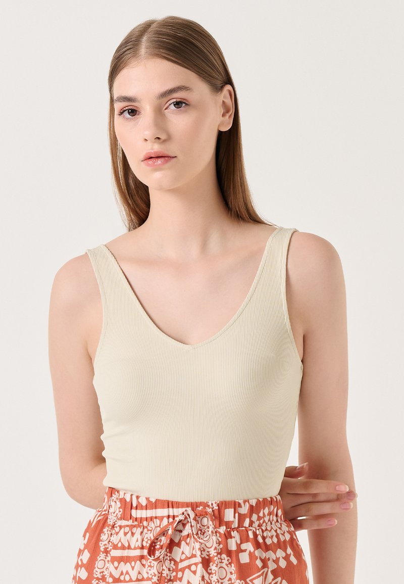Débardeur côtelé beige clair avec un décolleté en V. Le modèle l'a associé à une jupe orange à motifs comportant des motifs blancs.