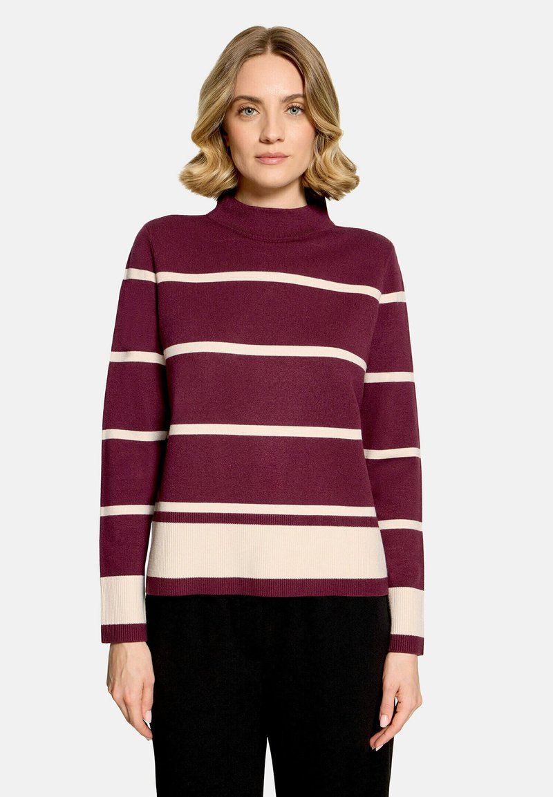 Femme portant un pull à col roulé rayé violet et beige à manches longues, debout devant un fond clair uni.