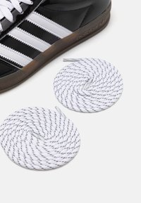 Svartvit Adidas-sneaker med glansigt material och tre vita ränder. Två vita skosnören med svart mönster är hoprullade.