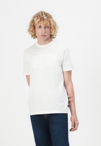 Osoba s plavom kovrčavom kosom, obučena u bijelu majicu s Guess logotipom i tamnoplave traperice, stoji nasuprot jednostavne bijele pozadine.