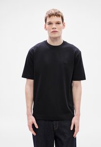 Fekete pamut póló kerek nyakkal és rövid ujjakkal, a mellkasán kis fekete logóval. Farmernadrággal párosítva.