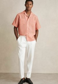 Rosa kortärmad skjorta med två bröstfickor, parat med vita skräddarsydda byxor och svarta loafers, bärs av en stående manlig modell.