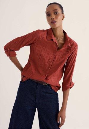 Femme portant une chemise boutonnée couleur rouille avec les manches retroussées et un pantalon ample large taille haute bleu foncé sur un fond uni.