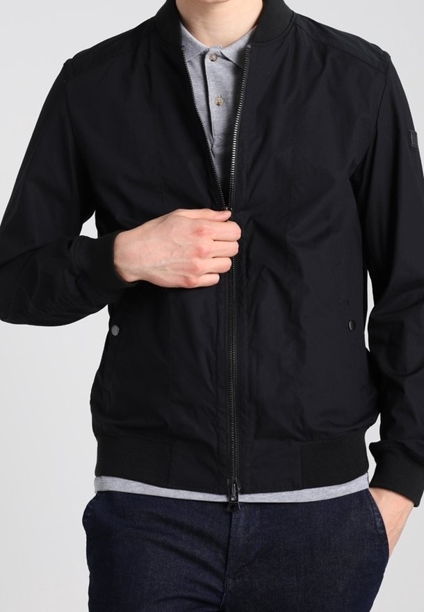 Homme portant une veste bomber noire zippée sur un polo gris, tirant la fermeture éclair vers le haut d'une main.