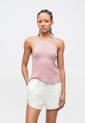 Mladá osoba s pletenými vlasmi, oblečená v ružovom rebrovanom tielku bez rukávov a bielych ľanových kraťasoch, stojaca na jednoduchom pozadí.