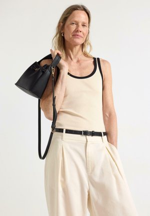 Kvinde iført en cremefarvet tanktop og brede cremefarvede bukser med sort bælte, der holder en sort håndtaske over skulderen mod en ensfarvet baggrund.