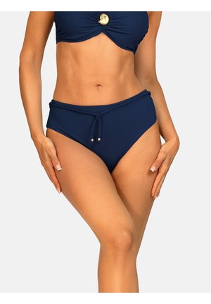 Kobieta ubrana w granatowe majtki od bikini z sznurkowym ściągaczem związanym z przodu oraz część dopasowanego topu z ozdobą w kształcie muszli.