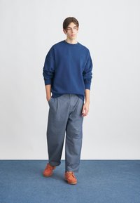 Navyblaues Sweatshirt mit Rundhalsausschnitt, lockere Passform; kombiniert mit hellgrauen, weit geschnittenen Hosen und braunen Schnürschuhen.