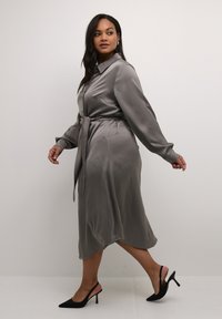 Graues Satin-Midikleid mit Kragen, langen Ärmeln, Taillengürtel zum Binden und fließender Silhouette, kombiniert mit schwarzen High Heels.