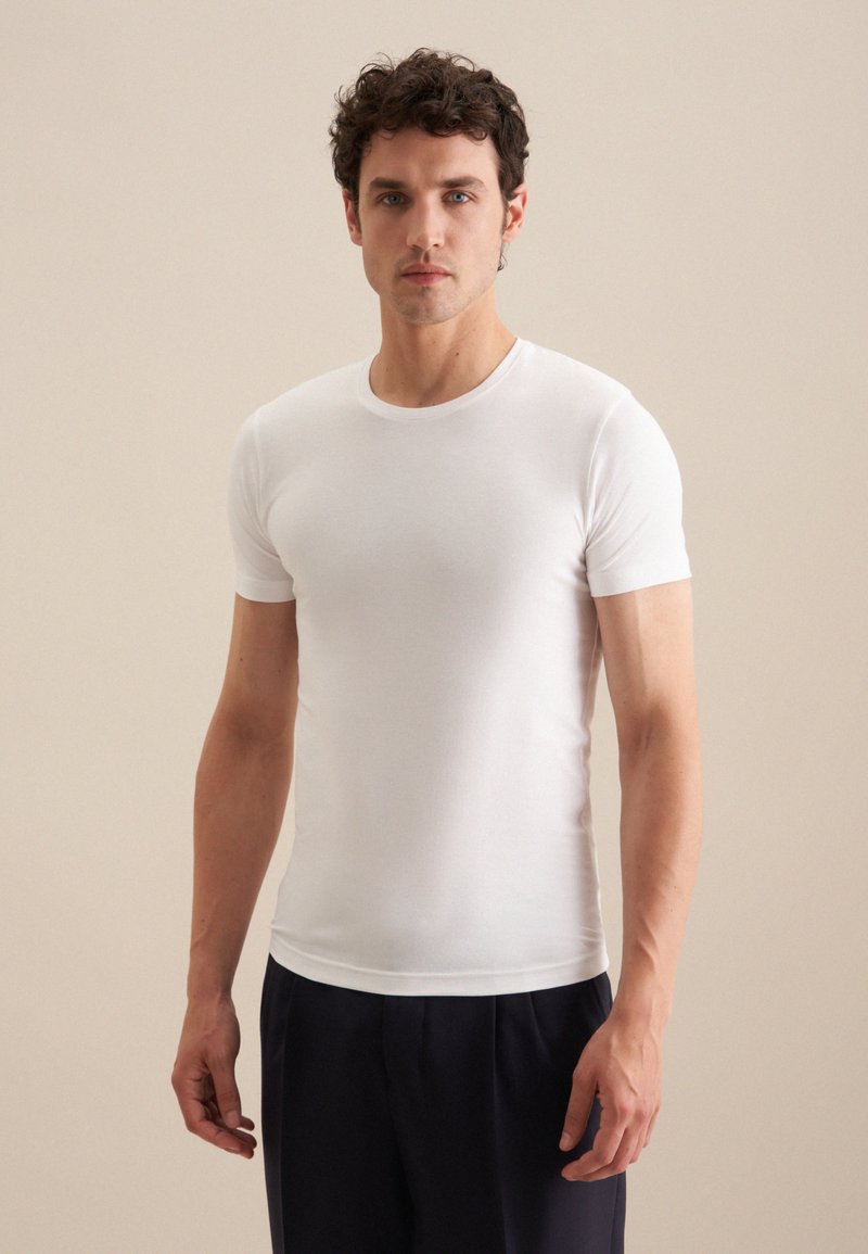 T-shirt blanc à manches courtes en tissu léger avec un col rond, une coupe ajustée et une texture lisse, associé à un pantalon sombre.