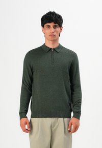 Tamsiai plaukti turintis vyras dėvi tamsiai žalią ilgomis rankovėmis polo megztinį su priekiniais užtrauktukais ir smėlio spalvos kelnes, stovintis priešais paprastą foną.