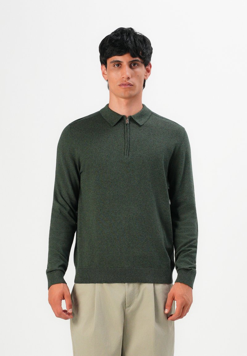 Tamsiai plaukti turintis vyras dėvi tamsiai žalią ilgomis rankovėmis polo megztinį su priekiniais užtrauktukais ir smėlio spalvos kelnes, stovintis priešais paprastą foną.