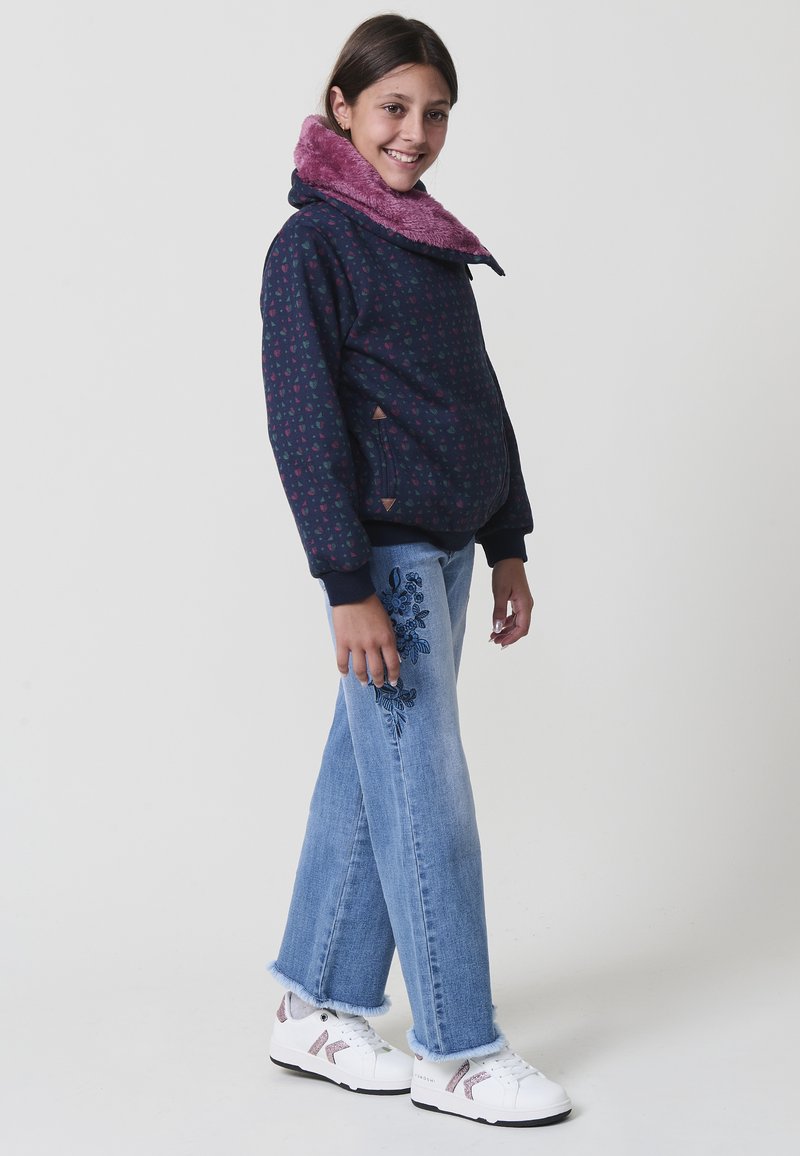 Ragazza sorridente in piedi, che indossa una giacca blu stampa con un colletto rosa peloso, jeans ricamati blu e sneaker bianche con accenti rosa.