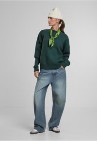 Grüner Sweatshirt, übergroße hellblaue Jeanshose, weiße Strickmütze, grüner gemusterter Schal um den Hals gebunden, lässiges Schuhwerk.