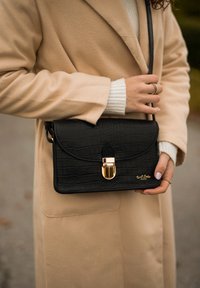 Schwarze Handtasche mit Krokodilmuster und goldener Schnalle, crossbody getragen. Der Griff hat eine strukturierte Oberfläche. Hellbeige Mantel im Hintergrund.
