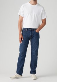 Hombre vestido con una camiseta blanca lisa, jeans azules y zapatillas blanco sucias, de pie con una mano en el bolsillo contra un fondo sencillo.