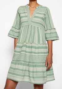 Grön mönstrad klänning med V-ringning, utsvängda ärmar och volangdetaljer. Har horisontella ränder och geometriska mönster över hela.