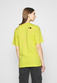 T-shirt di cotone giallo con maniche corte. Presenta un logo nero sulla parte superiore della schiena. Il tessuto ha una consistenza liscia e una vestibilità comoda.