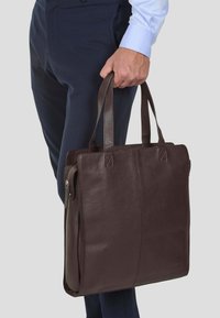 Borsa tote in pelle marrone con una texture liscia, forma rettangolare e due manici robusti. Presenta una tasca laterale con zip per ulteriore spazio di archiviazione.