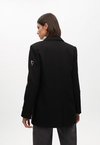 Cappotto nero sartoriale con colletto strutturato, dettagli decorativi a forma di cuore sulle maniche e bottoni decorativi sui polsini.