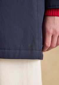 Mano appoggiata accanto all'orlo di una giacca blu navy con marchio "JOULES" discreto, polsino della manica rosso visibile e pantaloni color crema sotto.