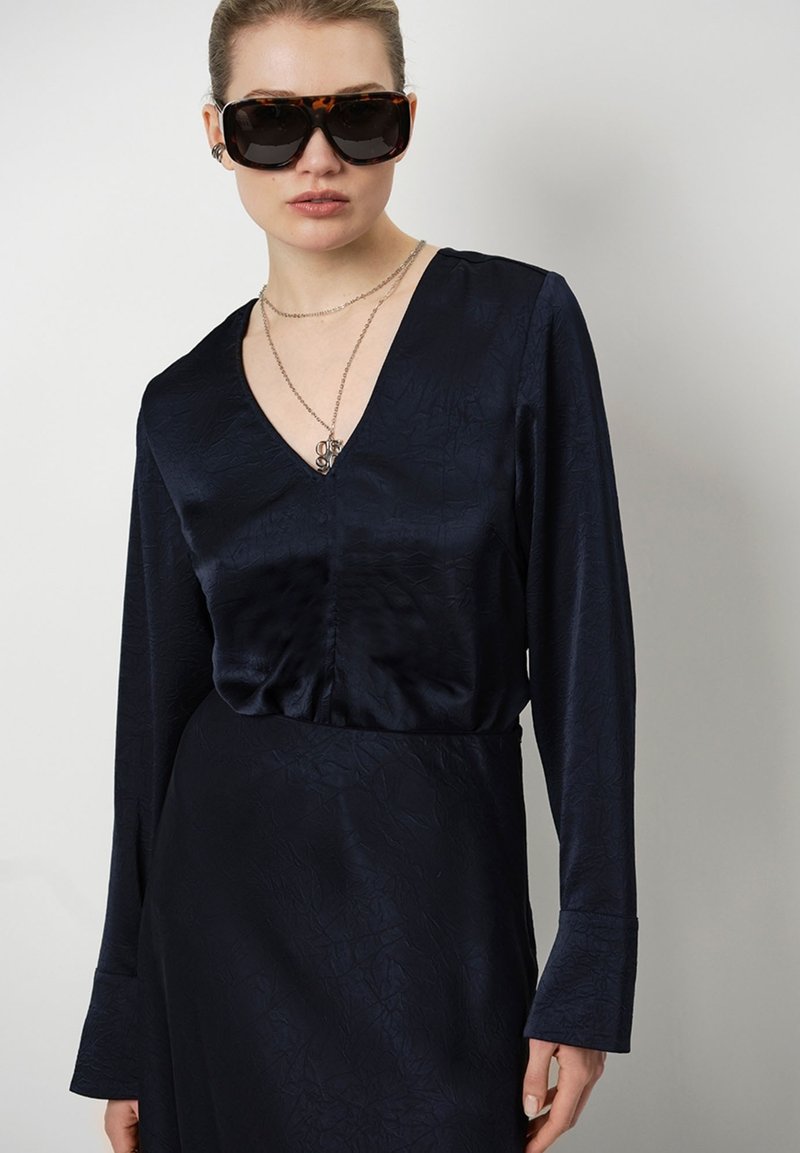 Marinblå långärmad klänning med v-ringning och texturerat tyg. Kombineras med stora, mörka sköldpaddsglasögon och lager av halsband.