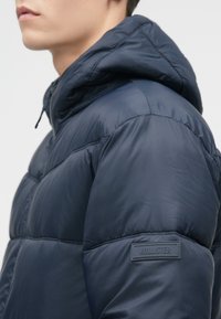 Chaqueta de plumas azul marino con capucha, con un acabado texturizado y un destacado logo de "Hollister" en la manga.