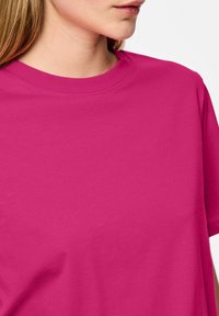 Jeune femme à la peau claire et aux cheveux châtain clair portant un t-shirt uni rose vif à manches courtes et col rond, sur fond blanc.