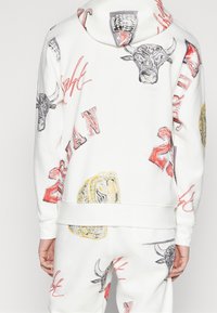 Sudadera blanca con estampados de cabeza de toro y logotipos en gris, rojo y amarillo. Textura suave, con capucha y puños acanalados.