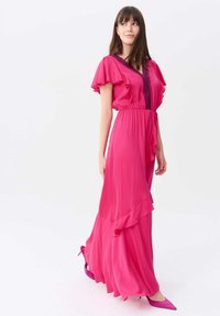 Maxikleid in lebhaftem Pink, mit Rüschenärmel, tailliertem Bund und geschichteter Saum. Ein Paar passende pinke High Heels rundet den Look ab.