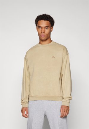 Sudadera beige con cuello redondo y mangas largas, que presenta un pequeño logotipo bordado en el pecho. Textura suave, corte relajado.