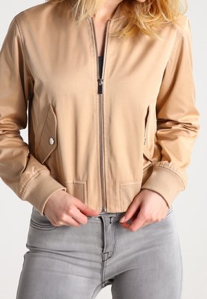 Femme portant une veste beige zippée avec des poches et un jean gris clair, mains ajustant la taille, regardant vers l'avant.