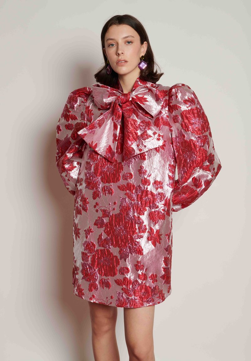 Λαμπερό ροζ φόρεμα με μεγάλους, φουσκωτούς ώμους και ένα εμφανές φιόγκο στον λαιμό, διακοσμημένο με κόκκινο λουλουδάτο μοτίβο.