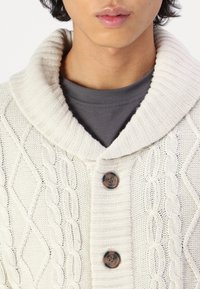 Krämfärgad stickad cardigan med hög, ribbad krage, kabelstickat mönster och två mörka knappar framtill.