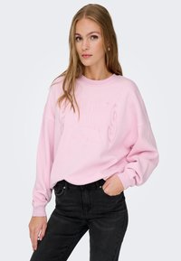 Sudadera de color rosa claro con el texto "PARIS" en relieve, con un corte holgado, puños acanalados y un cuello redondo, combinada con jeans negros.