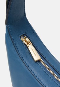 Gros plan sur un sac à main en cuir bleu avec une fermeture éclair dorée, une surface texturée et des coutures contrastées le long des bords.